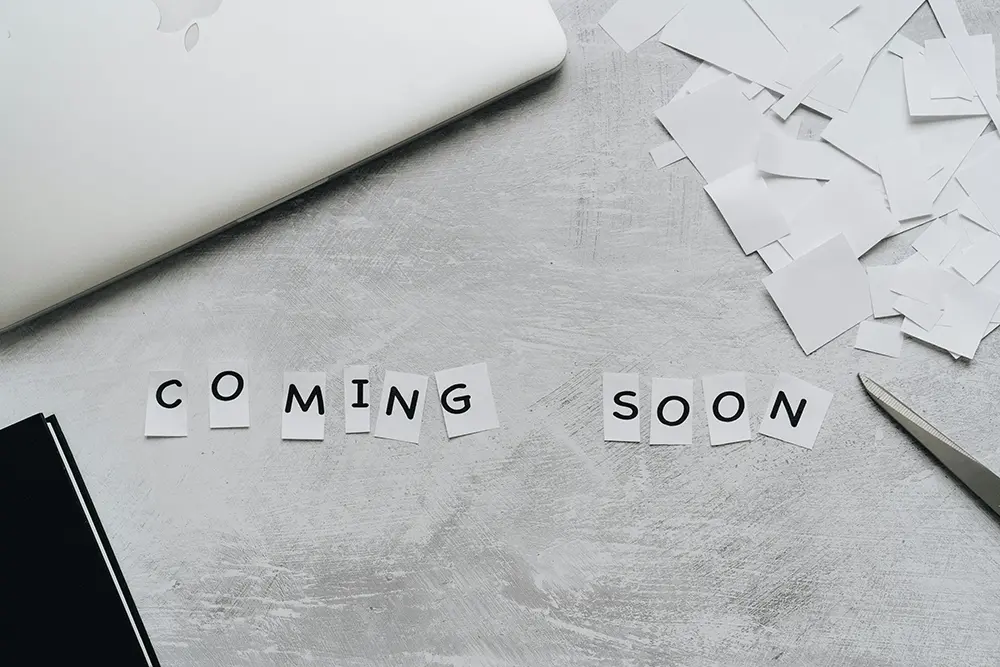 An image with the words "COMING SOON" written using cut-out letters on a grey background with torn paper around them.